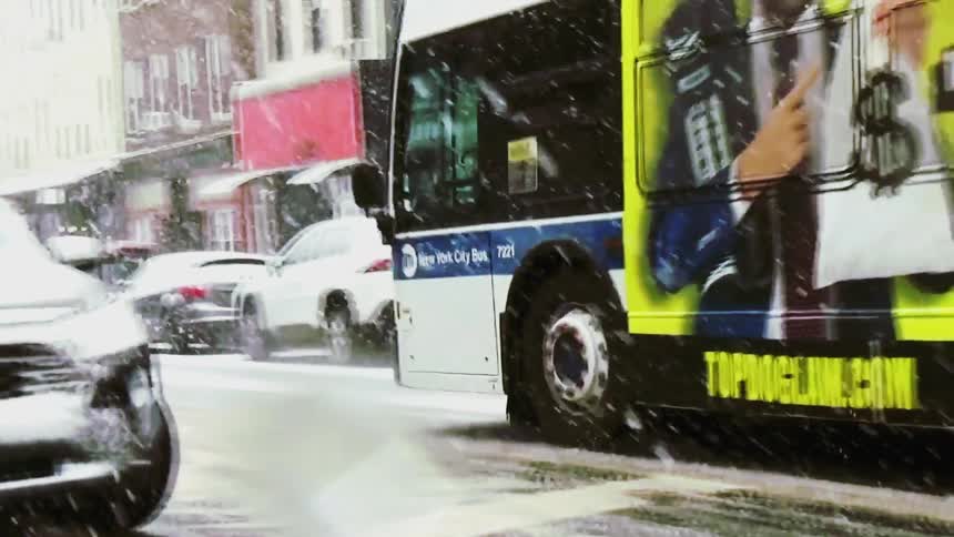 A city bus drives through a snowy urban street as traffic moves past apartment buildings during winter