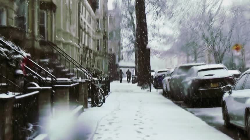 Two people walk away from the camera down a snowcovered sidewalk lined with brownstone buildings and parked carsheavy snow falls throughout the scene blurring the figures as they move toward a distant street corner