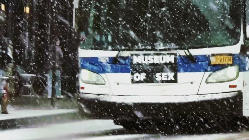 A person wearing red overalls crossing a street covered in snow while a bus with an advertisement on the side stops during a winter storm