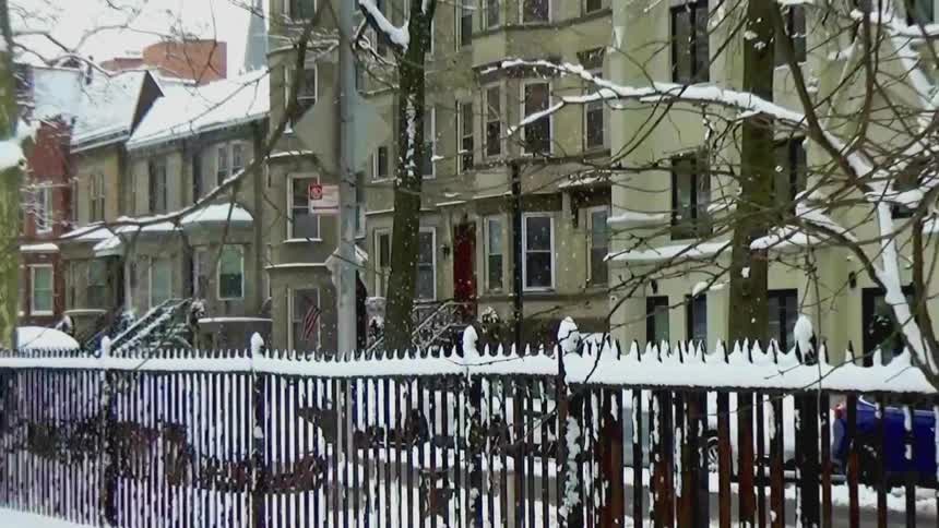 A snowy residential street lined with bare trees and row houses in winter daylight