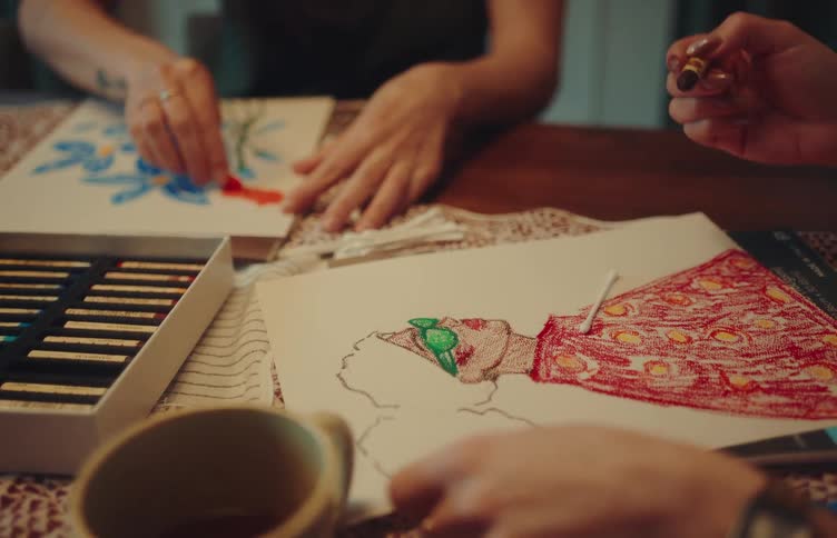 Two people at a table use colored pencils to create illustrations in a cozy indoor setting