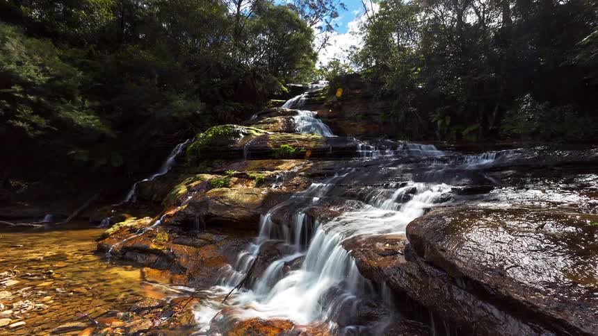 A cascading waterfall flows gently over wide flat rock tiers nestled within a lush green forest the sunlight filters through the trees creating bright reflections on the moving water and the shallow pool at the base