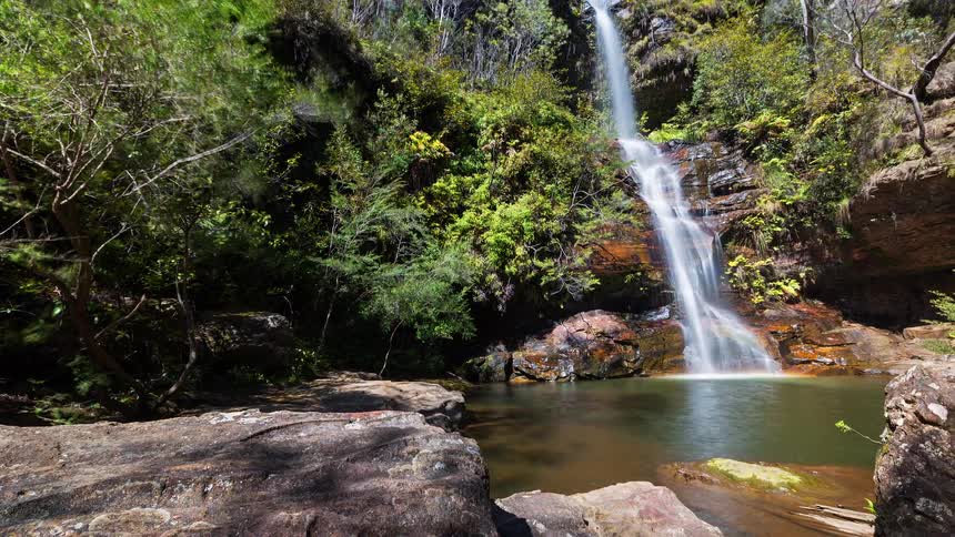 A waterfall flowing from a mountain into a pond with trees surrounding the area on a sunny day