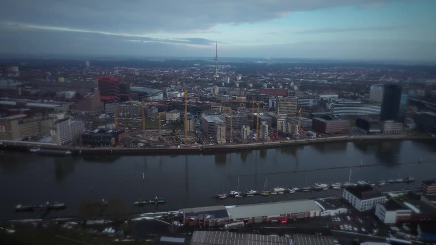 Aerial view of a city skyline along a river with cranes and buildings stretching under a cloudy sky
