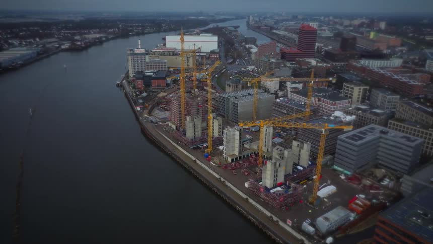 An aerial shot shows several yellow tower cranes working on a large building project along a wide river in a gray urban landscape during an overcast day as the camera slowly pans over the active construction site and nearby city blocks