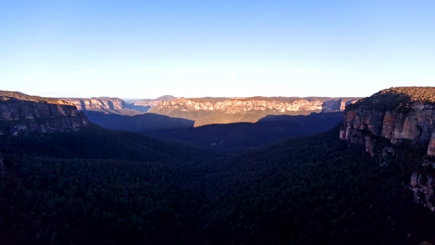 Cliffs overlook a forested mountain valley as shadows move across the landscape with the setting sun and stars glow in the night sky
