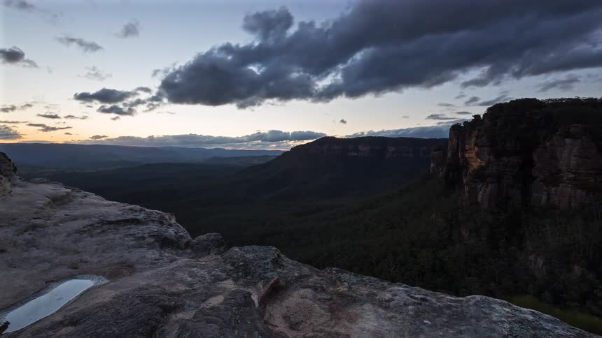 Clouds move over mountain rock formations and canyons as the sun sets on the horizon