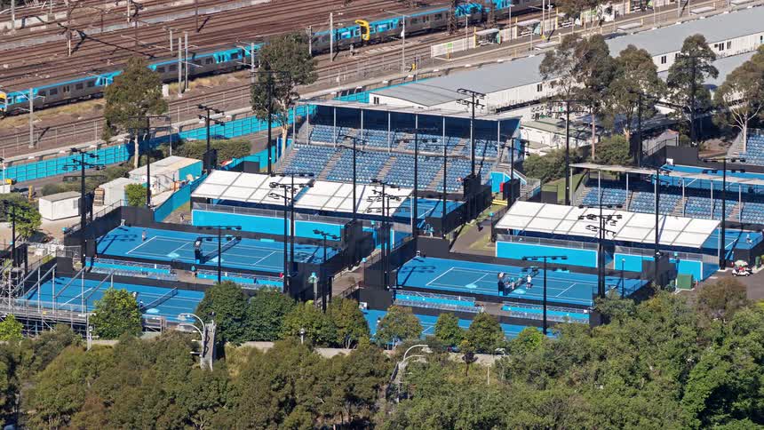 A busy tennis court at midday with a passing train visible on the left side and train tracks nearby