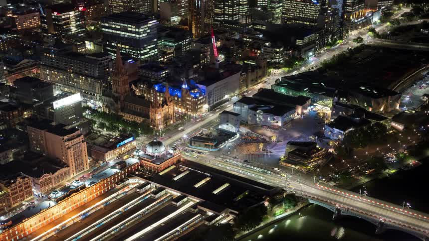 An aerial view shows the bright lights of a city at night with a large train station in the foreground while many cars and a tram move along the streets and a bridge over a river