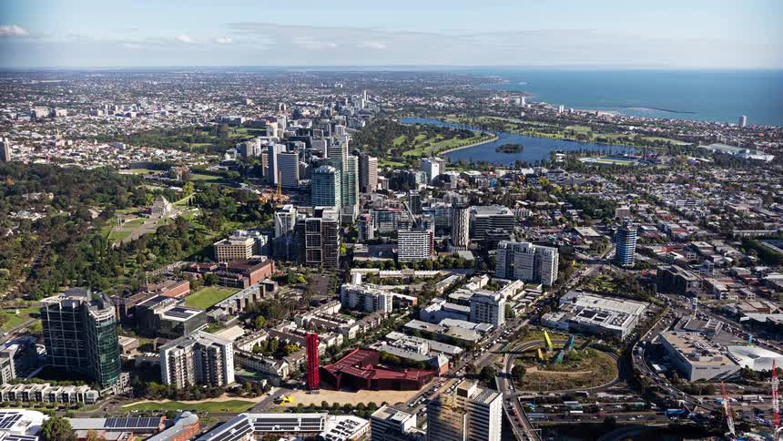 This aerial shot provides a sweeping view of melbournes skyline highlighting the contrast between the highrise buildings and the expansive greenery of albert park in the distance the calm blue waters of port phillip bay stretch toward the horizon und