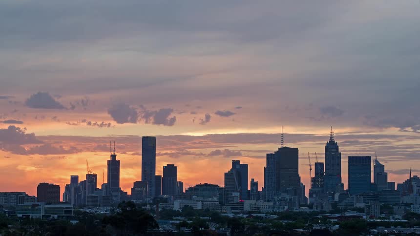 A view shows a city skyline at dusk with many buildings and houses as the sun sets and the sky turns orange and blue
