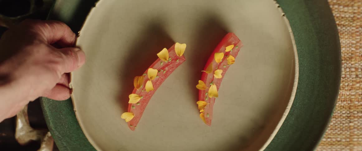 A hand lifting a ceramic plate with two cured fish slices garnished with yellow petals from a wooden table