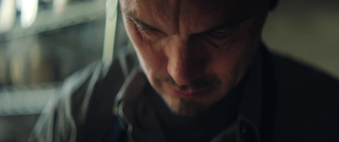 A middle aged man wearing a blue apron looks down with a concentrated and intense expression while working in a dimly lit indoor environment with metal shelves blurred in the background of the tight closeup shot