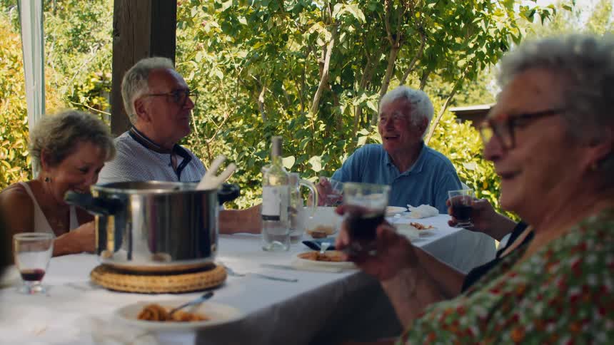 A group of elderly friends laughs and enjoys wine during an outdoor meal the sun filters through green leaves as they share a joyful moment around a table