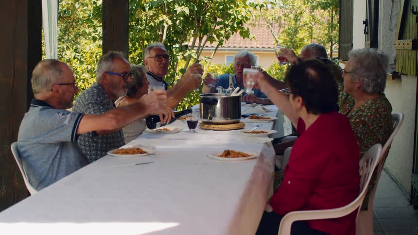 Small group of elderly men and woman toasting with wine and water glasses over a dinner table where a meal is served outdoors during day time
