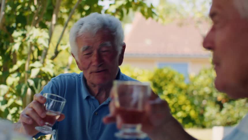 Two elderly men laugh and toast with drinks while enjoying a relaxed outdoor conversation