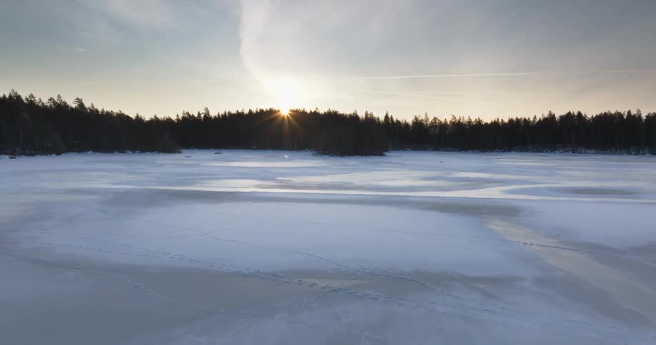 A view shows a frozen lake as the sun begins to set behind a line of trees in a forest during the winter