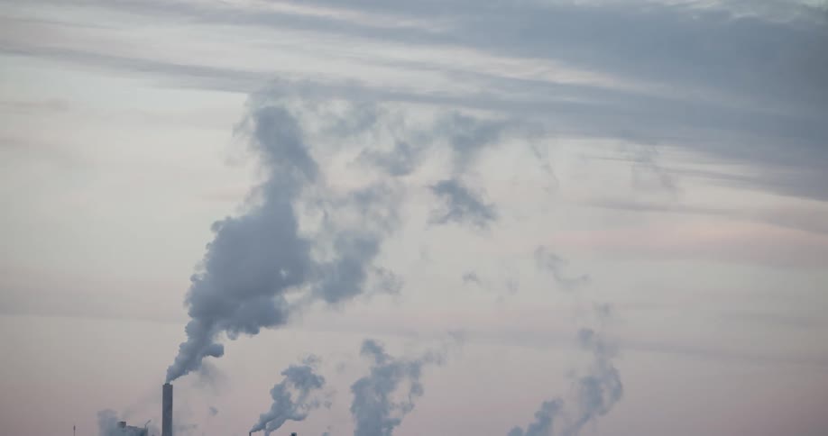A view shows many large chimneys as thick gray smoke rises from them into a cloudy sky above a factory during the day