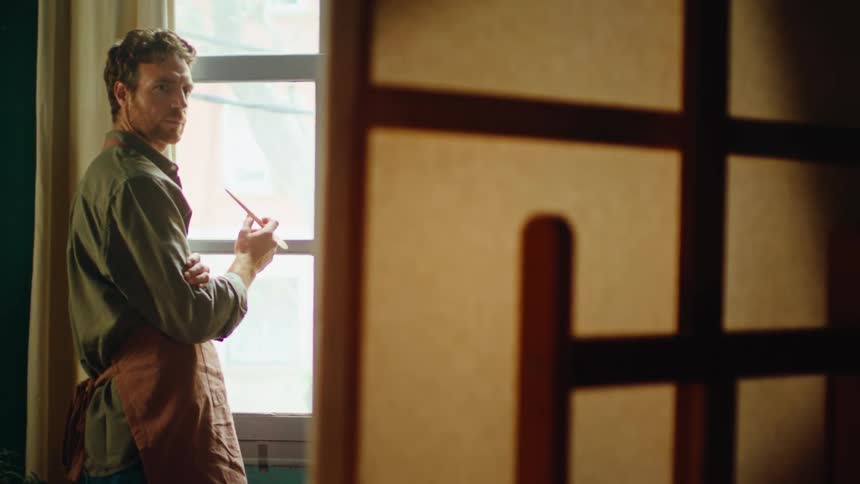 Painter standing near a window with natural light wearing an apron and holding an object in his hands