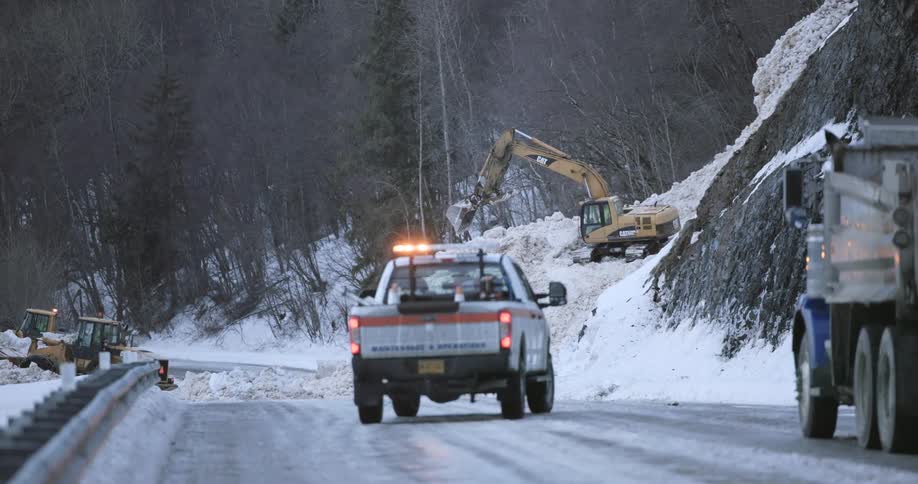 Heavy machinery and maintenance vehicles work to clear snow and debris from a mountain highway after a landslide or avalanche an excavator on the hillside moves earth while a maintenance truck with flashing lights monitors the icy road below