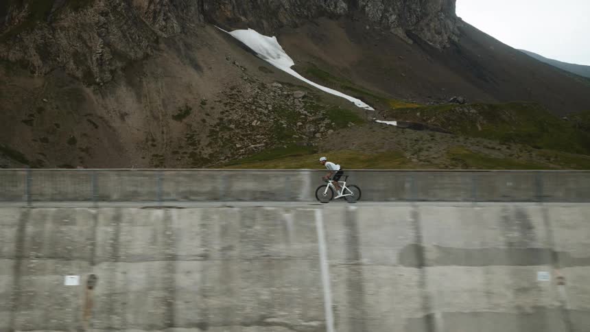 A view shows a person wearing a helmet and a white jersey as they ride a bicycle along a concrete wall next to a large body of water and a rocky mountain during the day