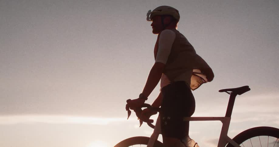 A view shows a person wearing a helmet and a white jersey as they stand with a bicycle while the sun sets behind them