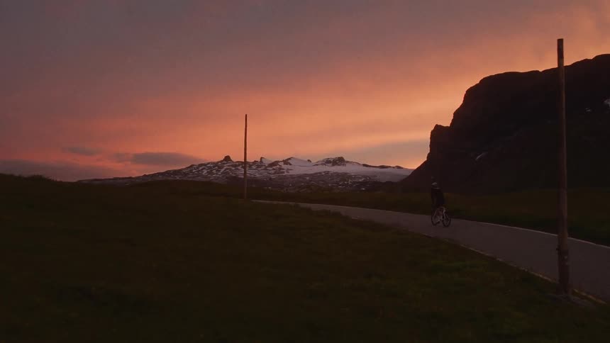 A lone cyclist rides along a winding road against a vibrant orange sunset and snowy mountain peaks