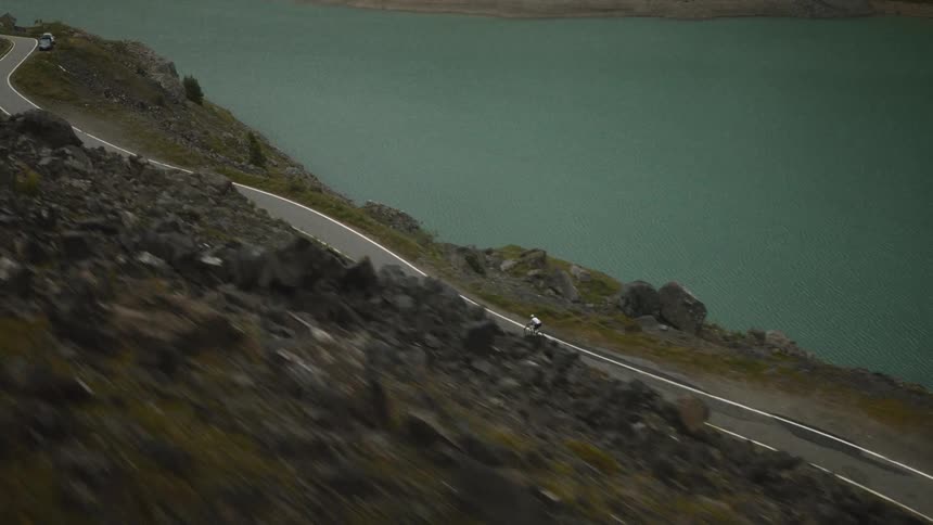 An aerial view shows a person as they ride a bicycle along a winding road next to a large body of water and a rocky hillside during the day