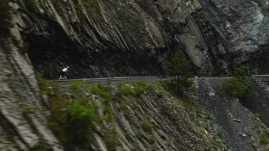 Cyclist rides along a narrow road beside a rocky cliff in a mountainous landscape