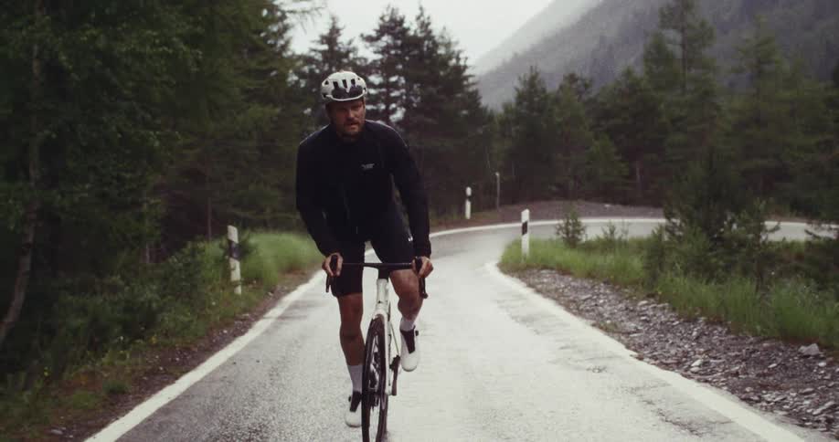 A view shows a person wearing a white helmet as they ride a bicycle along a road carved into a dark rocky cliffside during the day