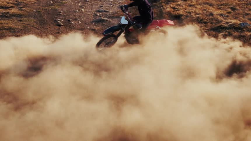 A view from above shows a person wearing a helmet as they ride a motorcycle along a dirt path on a brown hill while creating a large cloud of dust during the day