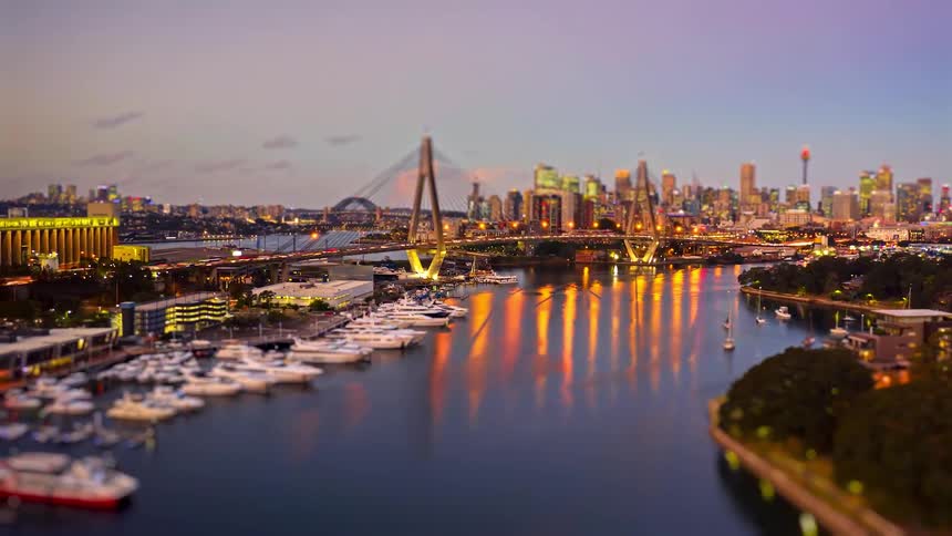An enchanting tiltshift aerial shot glides over a marina at dusk transforming the bustling city skyline and glowing bridge into a vibrant miniature world