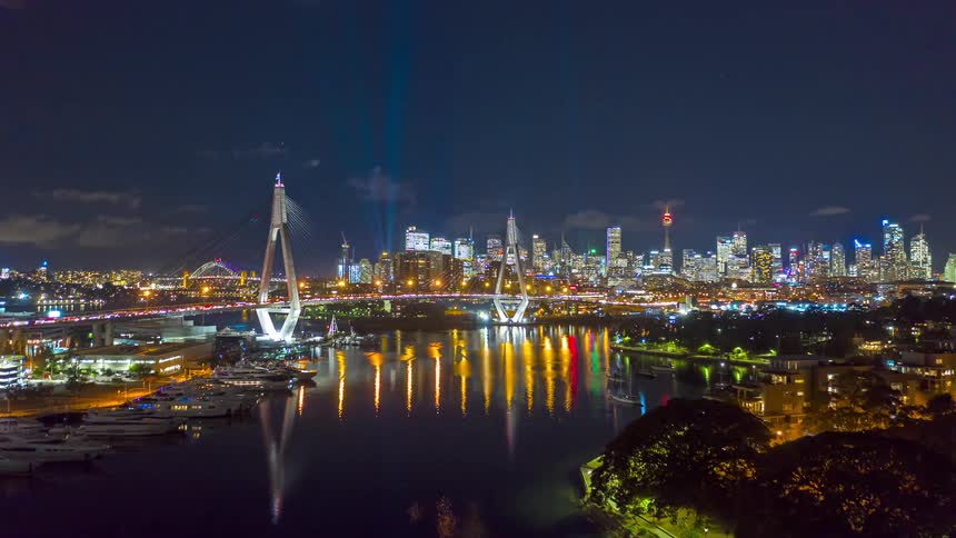A stunning aerial timelapse captures a vibrant city at night its illuminated bridge and skyline reflecting beautifully in the water below