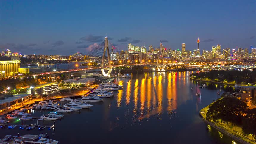 Illuminated cablestayed bridge crossing the river with a yacht marina on the left and an urban skyline reflected on the water at night
