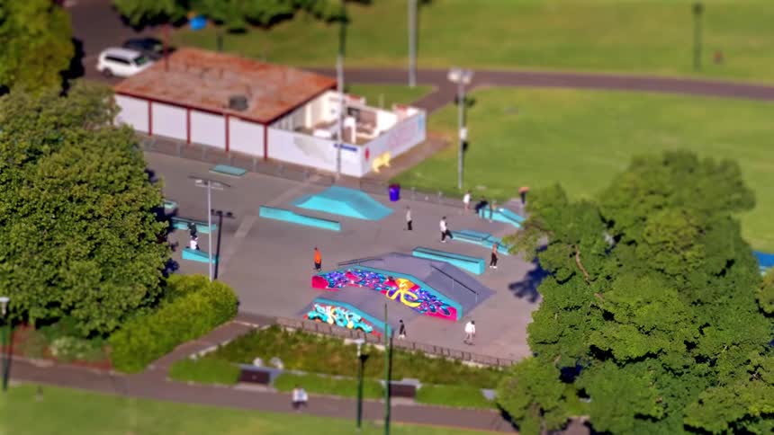 A small skateboarding park with colorful ramps and rails set within a green park where a few people are scattered around the concrete plaza