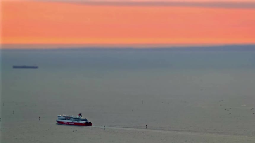 A red and white ferry glides across the water under a soft orange sunset sky