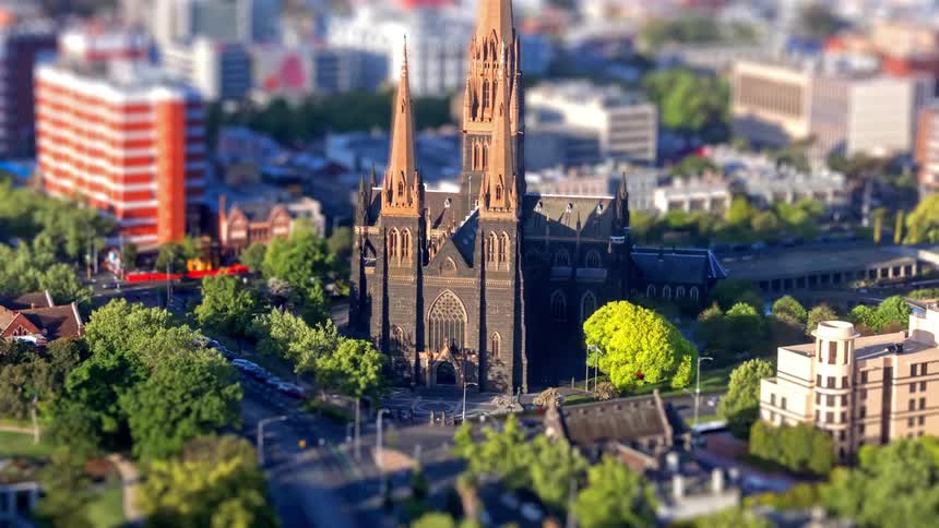 An aerial view of a giant cathedral surrounded by city streets buildings and green trees under bright daylight while cars passes by on a time lapse scene