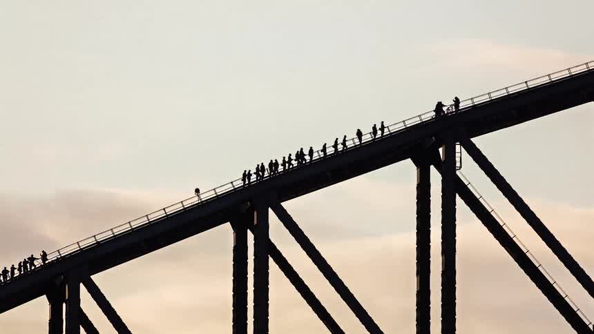 Silhouetted against a bright cloudy sky a singlefile line of figures slowly ascends the steep arched girder of a massive steel bridge during a scenic climb