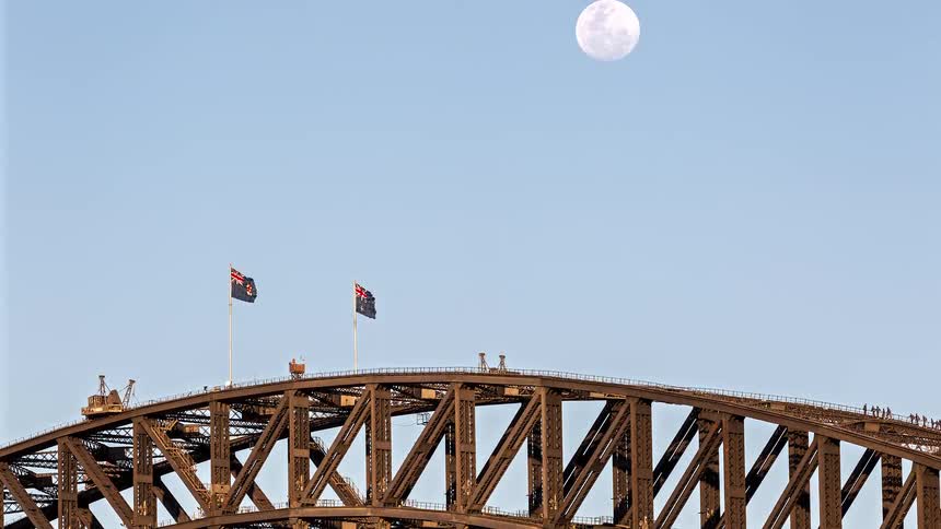 A view shows the top of a large metal bridge with two flags flying on it as a full moon shines in a clear blue sky during the day