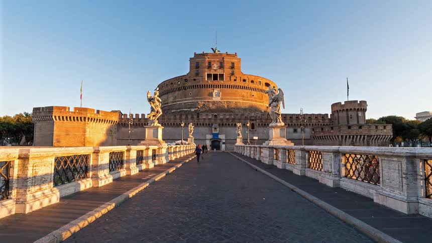 A historic stone fortress viewed from a pedestrian bridge as tourists walk toward it in clear daylight