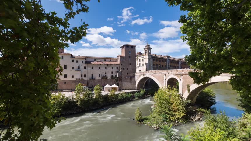 Ancient stone buildings and a bridge with arches stand along a flowing river under a bright blue sky with white clouds as green trees frame the view in the foreground