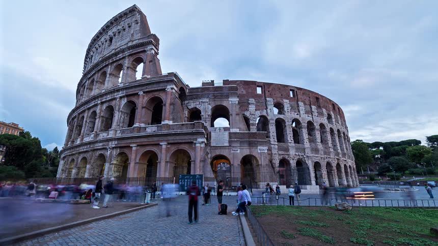 A view from a low angle shows the stone facade of the colosseum with its many arches as the sky transitions from day to night and warm lights begin to illuminate the ancient structure