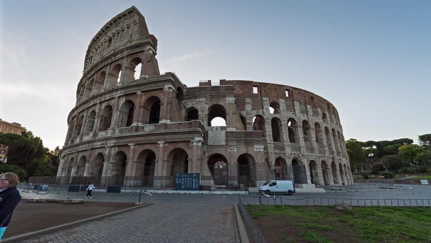 The roman colosseum stands under a clear blue sky as people walk along the surrounding paths