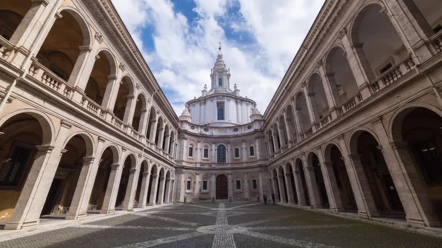 A symmetrical baroque courtyard with a spiral church dome under a cloudy sky