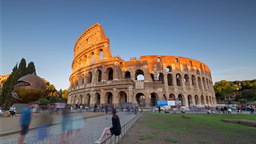 Visitors gather around the ancient roman colosseum as warm late day sunlight illuminates its iconic arches against a clear blue sky