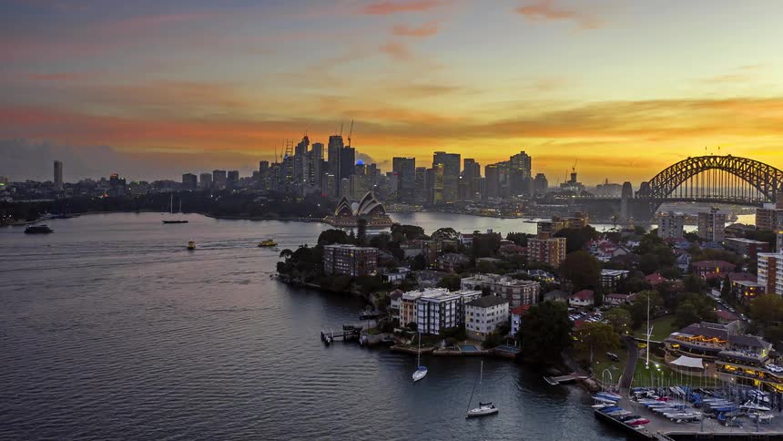 A high angle view shows the sydney city skyline and a large bridge over the harbor as the sun sets behind buildings creating a vibrant orange and yellow sky with silhouettes reflecting on the water