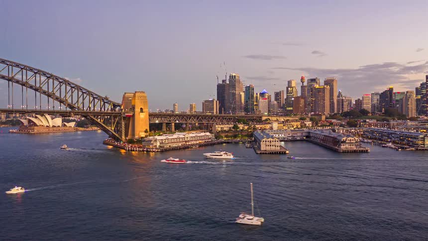 A view from the water shows a large steel bridge and city skyline with skyscrapers at sunset as boats move across the harbor during the late afternoon