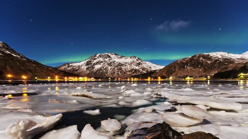 The northern lights glow above a snowcapped mountain and an icy harbor at night
