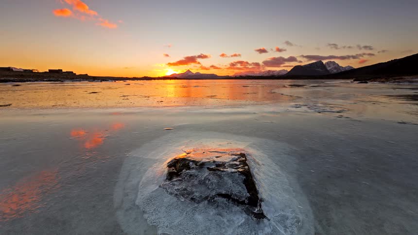 A fiery sunset reflects across a frozen lake where a lone rock breaks through the ice as distant mountains fade into the cold horizon
