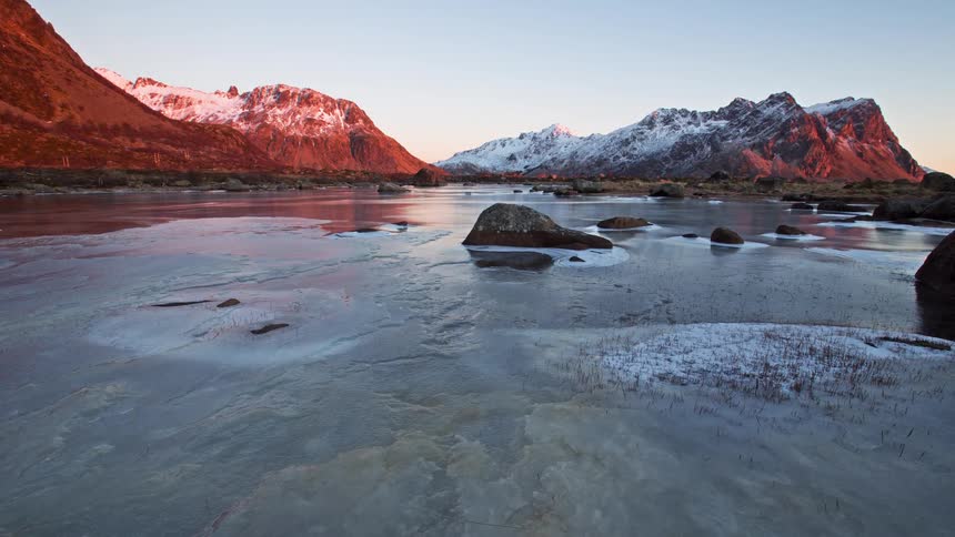 A frozen coastal inlet reflects snowcovered mountains glowing with warm sunrise light dotted with dark rocks embedded in the ice